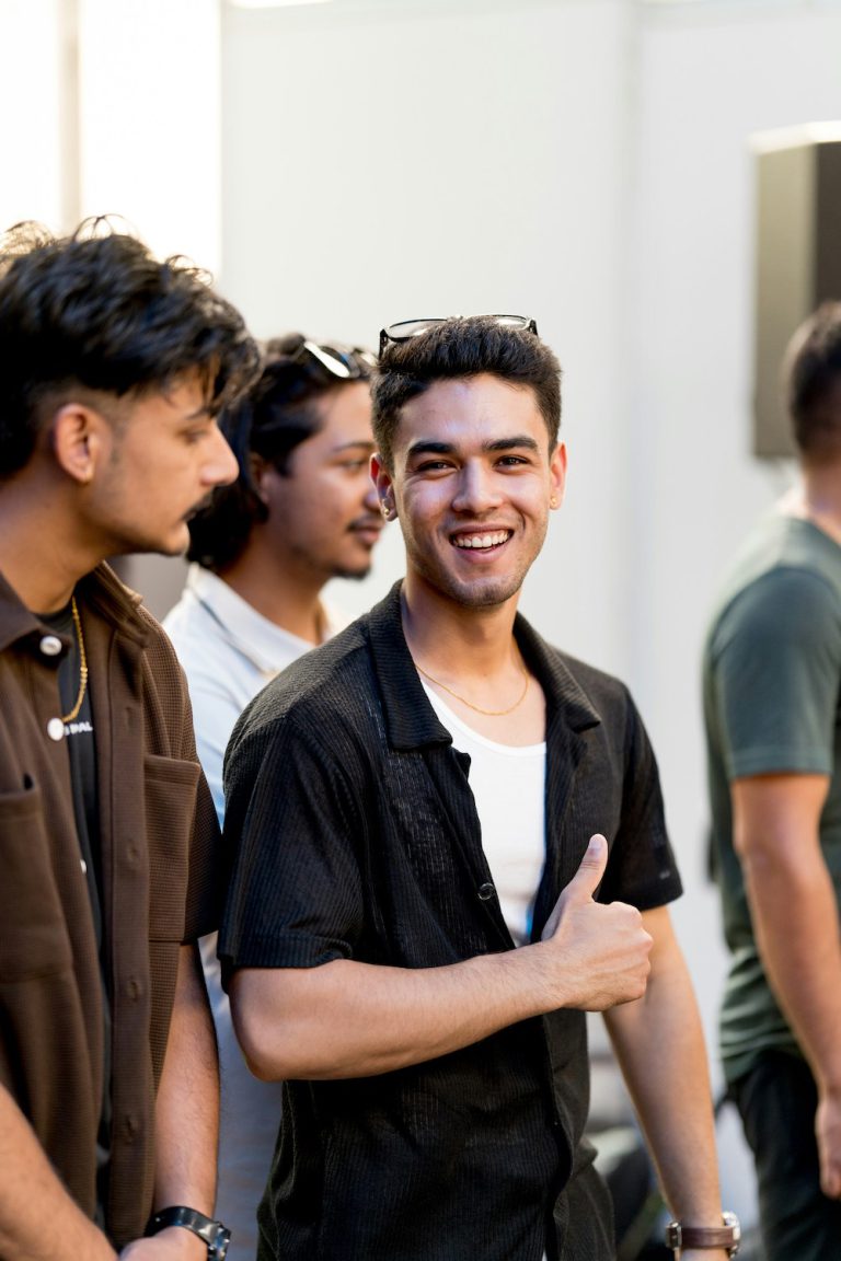 Young men hanging out. One of them is looking into the camera and smiling. He wears a black shirt open over a white undershirt.