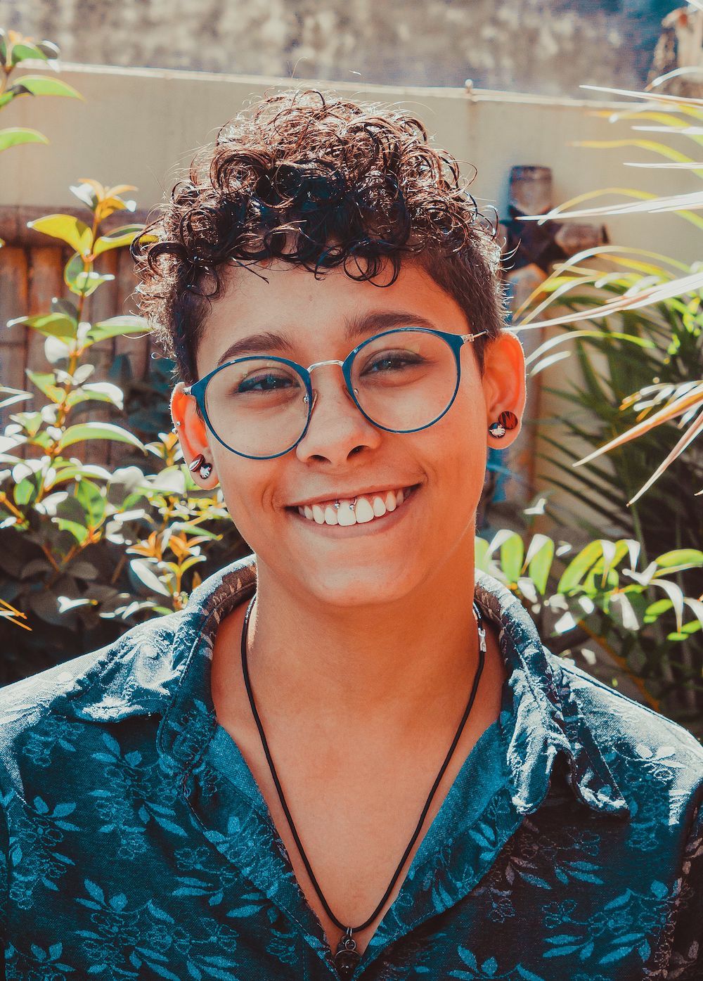Closeup pf young person with short curly hair glasses smiling directly at the camera in sunny light