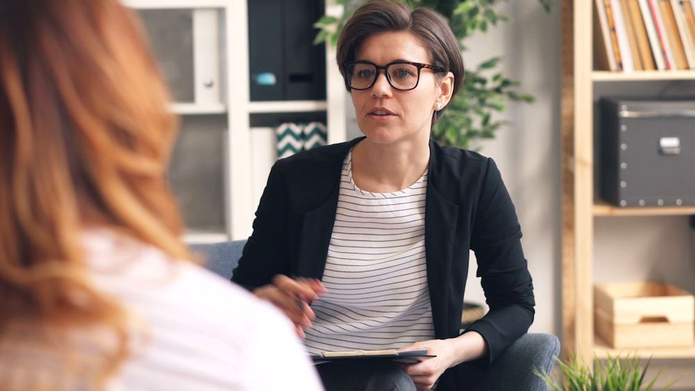 Closeup of female therapist with short hair and glasses talking to her client. She is wearing a striped shirt and black jacket.