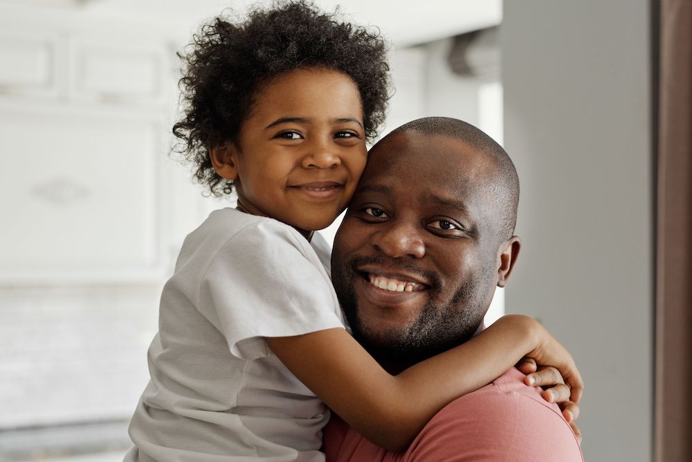 Closeup of black dad carrying his child in a living room with white paneling, both looking at camera and smiling.