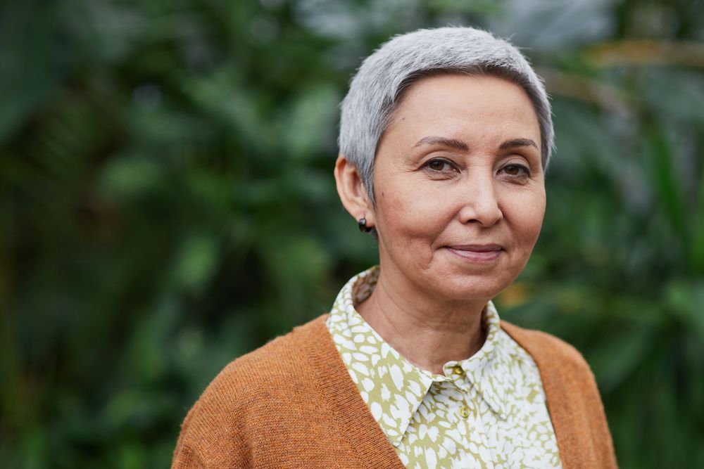 Older woman with cropped grey hair and orange sweater looking at the camara with a subtle smile