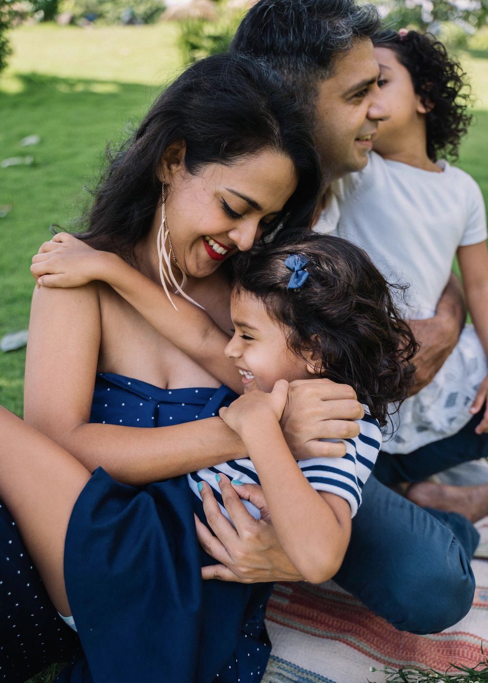 Closeup of hispanic family of 4 laughing in park. Closest is the mother in navy dress holding a small girl and laughing