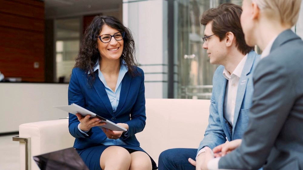 3 professionals in business suits talking together while sitting on a bench. Focus is on smiling hispanic woman with glasses, navy suit and long hair.