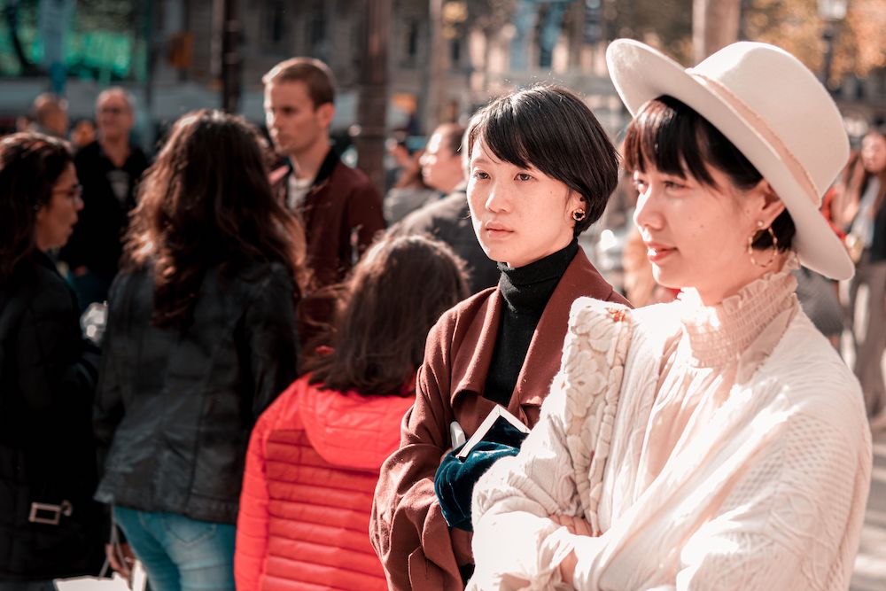2 middle aged asian women stand aside from a crowd and look on