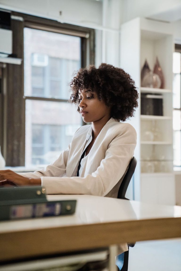 Young black female professional wearing a white blazer and natural hair typing on her laptop in an apartment full of light